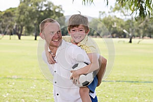 Young happy father carrying on his back excited 7 or 8 years old son playing together soccer football on city park garden posing s