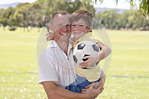 Young happy father carrying on his back excited 7 or 8 years old son playing together soccer football on city park garden posing s