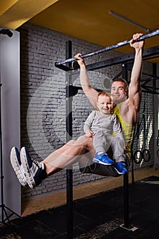Young happy father doing pull ups on the bar with son on his legs at the cross fit gym against brick wall.