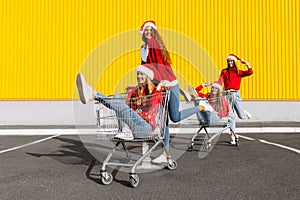 Young happy friends in Christmas sweaters and Santa Claus hats are having fun on a shopping trolley, near the mall, shopping