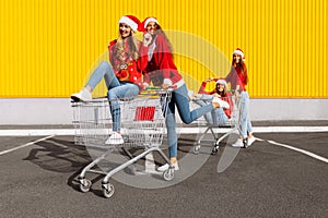 Young happy friends in Christmas sweaters and Santa Claus hats are having fun on a shopping trolley, near the mall, shopping