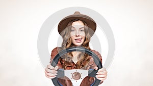 Young happy girl in a brown hat and leather jacket sticking out her tongue, holding a car steering wheel in her hands, on a light