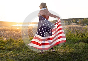 Young happy girl carelessly stands at sunset in the sun and holds the US flag behind her back