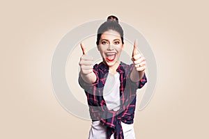 Young happy girl with casual style and bun hair thumbs up her finger, on beige blank wall with copy space looking at camera with t