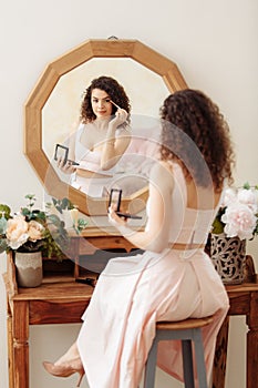 Young happy girl with curly hair does makeup in front of a vintage mirror. A beautiful woman in a pink dress paints her eyes