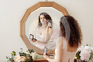 Young happy girl with curly hair does makeup in front of a vintage mirror. A beautiful woman in a pink dress paints her eyes