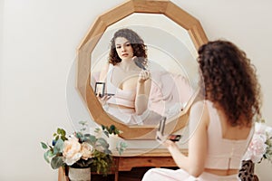 Young happy girl with curly hair does makeup in front of a vintage mirror. A beautiful woman in a pink dress paints her eyes