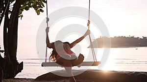 Young happy girl swaying on rope swings near amazing beach of paradise island at sunset