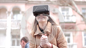 Young happy girl wearing vr virtual reality headset glasses having fun playing outside on street in beige outwear coat