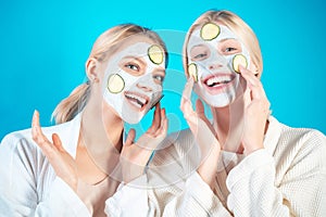 Young happy girls do facial mask with slices of cucumber at blue background. Skin care and treatment, spa, natural