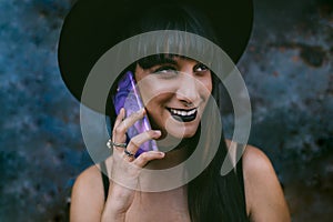 Young happy Halloween witch woman using smart phone. Beauty Witch lady with black mouth in the darkness, wearing a witch hat.