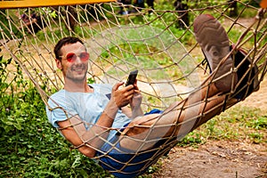 Young Happy Handsome Man Smiling In Hammock With Smartphone.