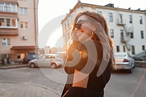 Young happy hipster woman with a positive smile in sunglasses in a stylish T-shirt in a coat in the city near houses