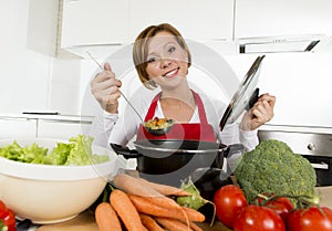 Young happy home cook woman in red apron at domestic kitchen holding saucepan tasting hot soup