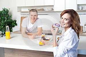 Young happy husband in the kitchen care for his wife. Woman eating donut and drinking juice. Lovestory, couple in love photo
