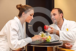 Young happy just married Caucasian couple in white bathrobes having fruits after spa on honeymoon. Man offers an apple to the woma