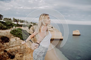 Young happy lady is sitting on a high and raising her arms up with panorama view on ocean coastline