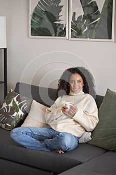 Young happy latin woman drinking tea or coffee relaxing at home on couch.