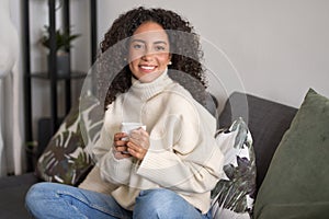 Young happy latin woman drinking tea relaxing at home sitting on couch.