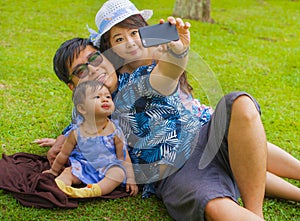Young happy loving Asian Chinese family with parents and sweet baby daughter at city park together with father taking selfie pic