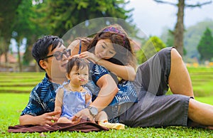 Young happy and loving Asian Chinese parents couple enjoying together with sweet daughter baby girl sitting on grass at green city