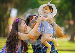 Young happy and loving Asian Chinese parents couple enjoying together with sweet daughter baby girl sitting on grass at green city