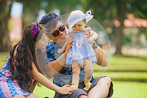 Young happy and loving Asian Chinese parents couple enjoying together with sweet daughter baby girl sitting on grass at green city