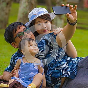Young happy loving Asian Japanese family with parents and sweet baby daughter at city park together with father taking selfie pic