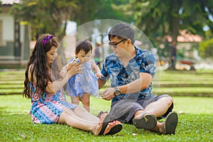 Young happy and loving Asian Japanese parents couple enjoying together with sweet daughter baby girl sitting on grass at green cit