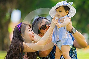 Young happy loving Asian Japanese parents couple enjoying together sweet daughter baby girl sitting on grass at green city park in