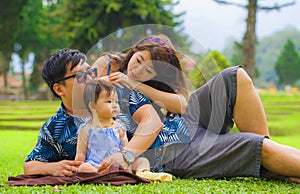 Young happy loving Asian Japanese parents couple enjoying together sweet daughter baby girl sitting on grass at green city park in