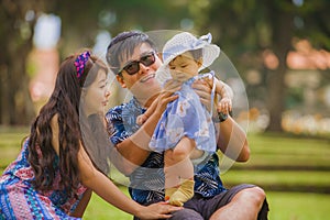 Young happy loving Asian Japanese parents couple enjoying together sweet daughter baby girl sitting on grass at green city park in