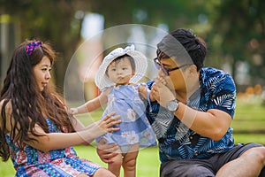 Young happy loving Asian Japanese parents couple enjoying together sweet daughter baby girl sitting on grass at green city park in