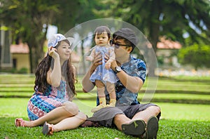 Young happy loving Asian Japanese parents couple enjoying together sweet daughter baby girl sitting on grass at green city park in