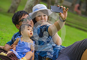 Young happy loving Asian Korean family with parents and sweet baby daughter at city park together with father taking selfie pic