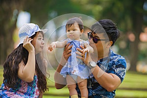 Young happy loving Asian Korean parents couple enjoying together sweet daughter baby girl sitting on grass at green city park in