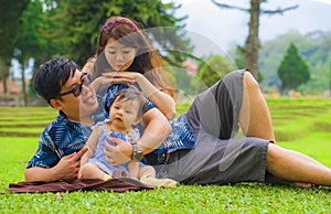 Young happy loving Asian Korean parents couple enjoying together sweet daughter baby girl sitting on grass at green city park in