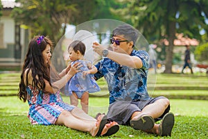 Young happy loving Asian Korean parents couple enjoying together sweet daughter baby girl sitting on grass at green city park in