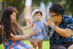 Young happy loving Asian Korean parents couple enjoying together sweet daughter baby girl sitting on grass at green city park in