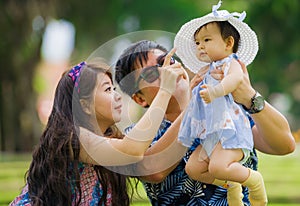 Young happy loving Asian Korean parents couple enjoying together sweet daughter baby girl sitting on grass at green city park in