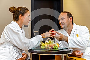 Young happy loving Caucasian couple of man and woman in white bathrobes talking after spa on honeymoon. Different fruits lying on