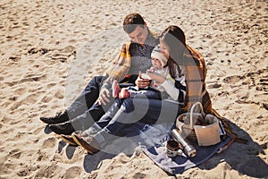 Young happy loving family with small child at picnic, enjoying time at beach sitting and hugging near ocean, happy lifestyle famil