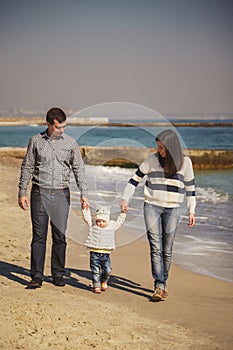 Young happy loving family with small kid in the middle, enjoying time at beach walking near ocean, holding arms, happy lifestyle f