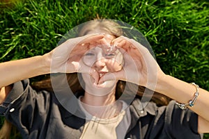 Young happy loving woman showing heart sign, looking at camera lying on grass