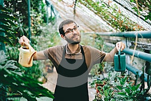 Young happy man gardener environmentalist holding a watering cans in hands, caring for plants in greenhouse