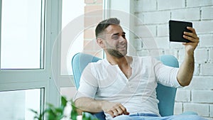 Young happy man have online video chat using digital tablet computer sitting on balcony in modern loft apartment