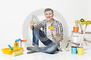 Young happy man holds bundle of dollars, cash money, cretit card, sits on floor with instruments for renovation