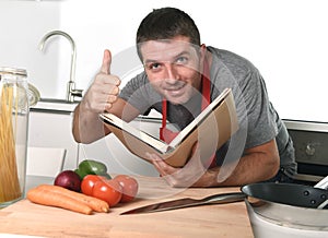 Young happy man at kitchen reading recipe book in apron learning cooking