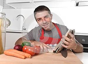 Young happy man at kitchen reading recipe book in apron learning cooking