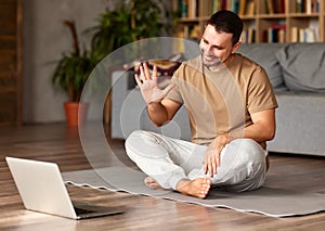 Young happy man in lotus pose waving at webcam on laptop while teaching yoga online photo